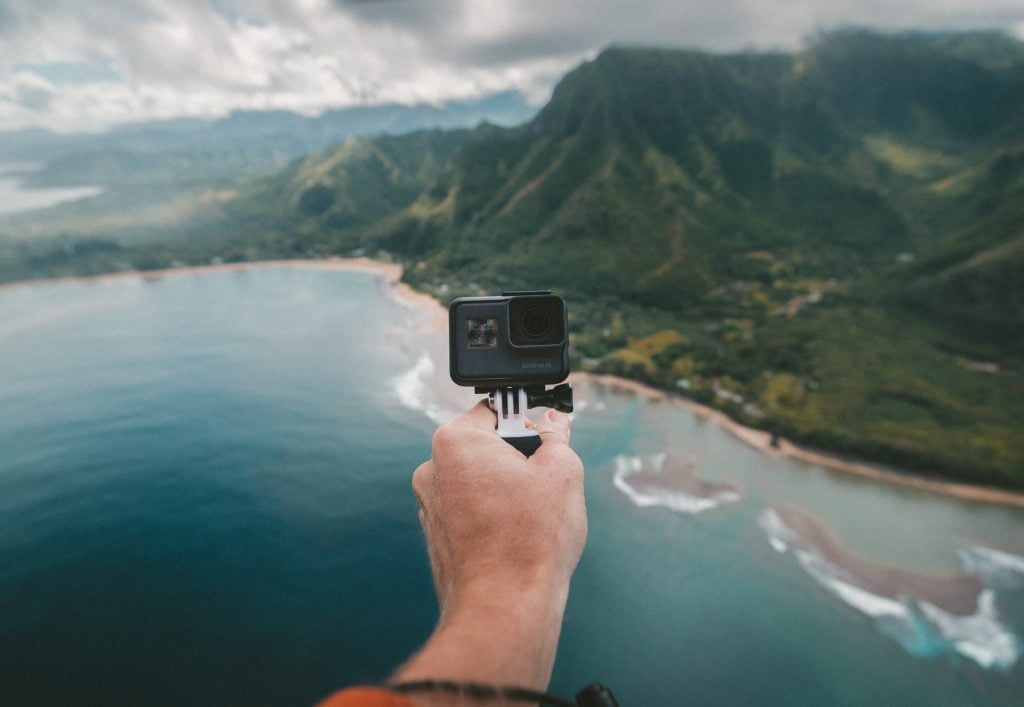 Uma mão segurando a melhor GoPro com uma paisagem de praia e montanha ao fundo.